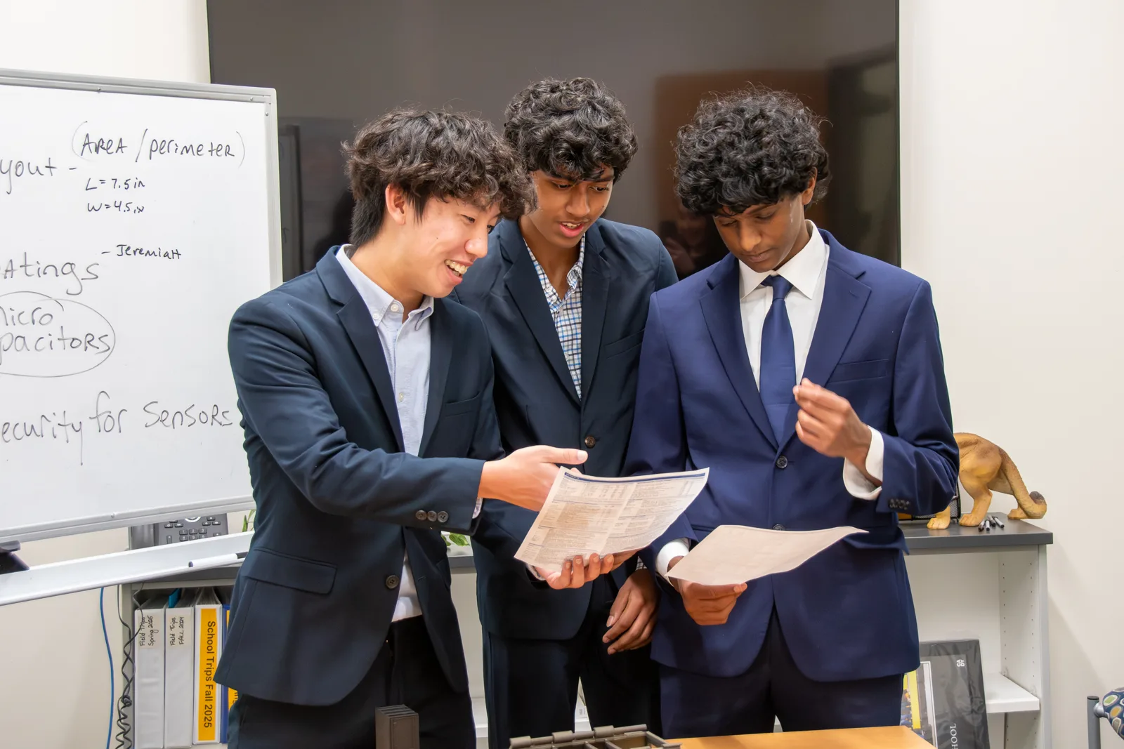 Three Apex High seniors in suits read printed design notes together beside a whiteboard covered in notes about panel layout and sensors.